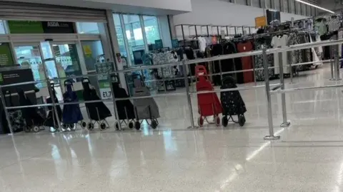 Aga Moss A row of shopping trolleys lined up next to railings inside an Asda supermarket, with dresses on railings in the background