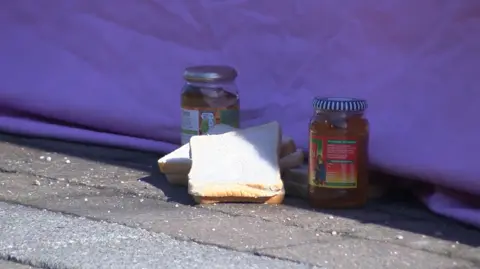 Two sandwiches made with white bread and two jars of marmalade on a paved ground, with the wall of a purple tent behind them.