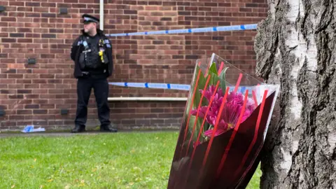 Stuart Woodward/BBC A bouquet of flowers left by a tree. In the photo there is also a police officer stood by a police cordon. 