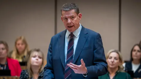 Senedd Cymru Rhun ap Iorwerth is stood in the Senedd with a group of Plaid MSs behind him, wearing a suit.