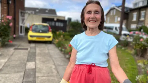 Aisha Iqbal / BBC An older lady standing in her front garden and driveway. She wears gardening gloves. There is a car parked behind her and there are flowers and grass verges around her which look pretty and well kept.