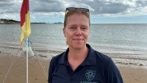 Jodie Halford/BBC A woman wearing a blue polo shirt smiles at the camera. Behind her is the beach and shoreline, and a red and yellow flag on a stand.