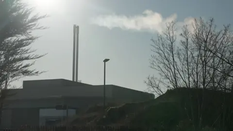 The Beddington Energy Recovery Facility is seen from outside, with two tall chimneys releasing a plume of steam into the sky, surrounded by trees and fencing.