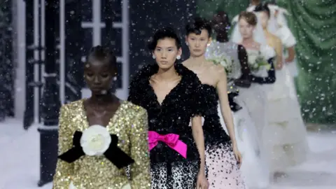 Isabel Infantes/Reuters Seven models stand in a line on a snow-covered stage and snow is falling on them. They are wearing glitzy dresses