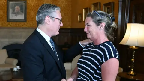 The Prime Minister in a dark blue suit shakes hands with Cheryl KOrbel who has black dress with white thin hoops