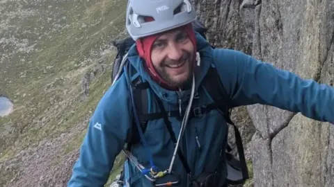 West Yorkshire Police A picture of a bearded man in climbing gear, positioned next to a wall of rock. He wears a grey helmet and blue sweater. He smiles at the camera.