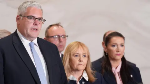 A group of politicians stand behind a podium at a press conference, with one man addressing microphones while others look on.