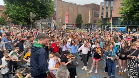 BBC Hundreds of musicians young and old stand in a city centre park, playing a variety of traditional instruments as they are guided by one man in the foreground.