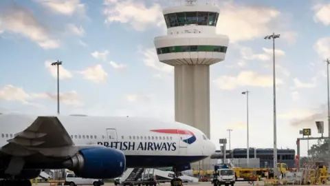 A British Airways aircraft parked at an airport stand. A tall, white control tower is in the background.