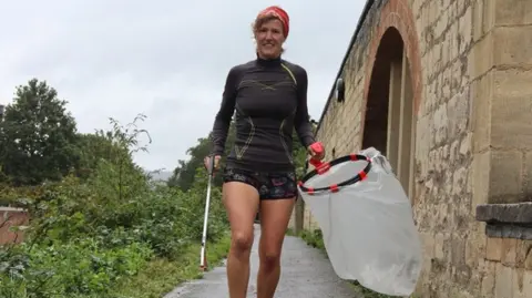 Councillor Gill Thomas wearing a long black running top and black shorts. She is running down a path whilst holding a litter picker in one hand and an empty plastic bag in the other.