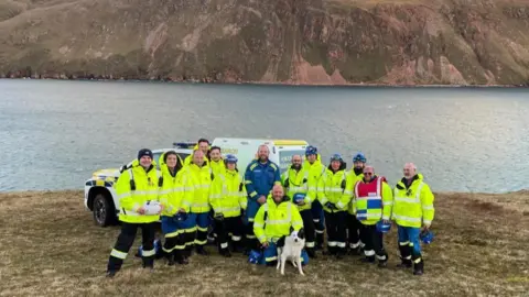 HM Coastguard About a dozen Coastguard team members are photographed on a clifftop with black and white collie dog Whisp, with sea and cliffs in the background, they are smiling at the camera after a successful rescue operation.