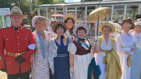 A group of eight people wearing regency clothing. The women are wearing long modest dresses with short lace gloves, and one is holding a yellow frilled parasol above her head. There is also a man linking arms with one of the women on the left, wearing a red army parade tunic with gold detailing. They are all standing on the train platform at Bath Spa station on a sunny day.