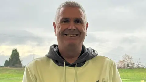 Darren Prosser Darren is wearing a cream hooded top with a grey hood. He is smiling at the camera and has cropped grey hair.