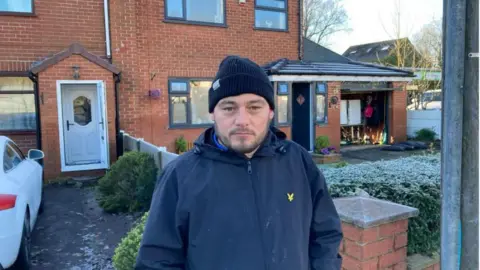 A man stands outside his front door, wearing a coat and a wool hat 