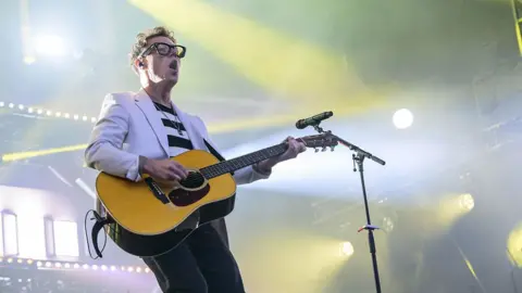 Getty Images Callum Beattie is playing guitar on stage. The picture has been taken from below, looking up at him. He is wearing thick-rimmed, black glasses, a black and white striped T-shirt, jeans and a white suit jacket. There are yellow and white stage lights illuminating the picture