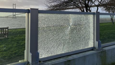 A damaged glass panel on the Hessle tidal flood defence. The Humber Bridge can be seen in the background.