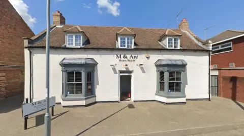Google The outside of M & Arc Hair and Beauty, with its sign in black letters above the door. It is an old detached property that has two bay windows on the ground floor and three small ones at the top. It is painted white and there is an advertising billboard on the driveway.