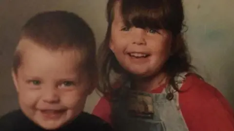 Undated photograph of James and Kelly as youngsters. James has blond short hair and is wearing a black top. Kelly has long dark brown hair with a long fringe. She is wearing a red top and denim dungarees. They are smiling.