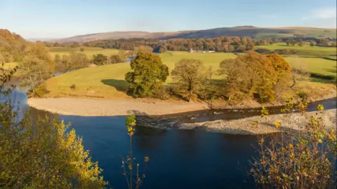 A river in front of a pebbled river bank and green fields. Hills can be seen in the background alongside trees. The leaves on the trees are just turning orange.