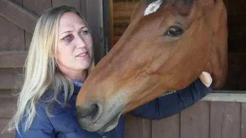 Becci stood next to a stable door with Barney - a brown horse - sticking his head out. Becci, who has long blonde hair and is wearing a blue coat, has a black eye from when she was knocked off Barney. 