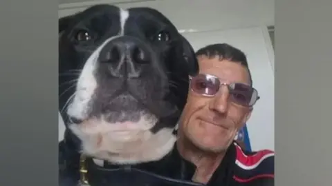 A close-up photo of a black and white dog's head and a man wearing glasses. Both are looking into the camera