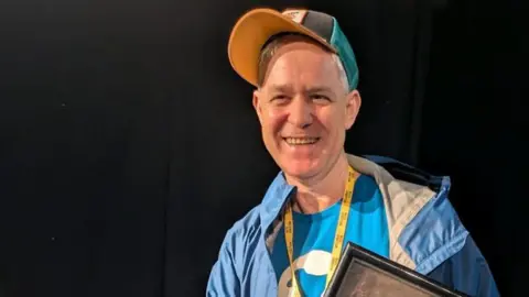 BBC A man with grey hair, wearing a blue baseball cap, blue t-shirt and blue jacket, holding a black framed item, standing against a black background.