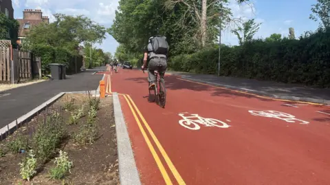 General view of Adams Road, which is a red road with double yellow lines, cycles painted on to the road and a cyclist on it.
