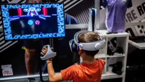 A young child has short brown hair and is wearing a orange t-shirt as he has a virtual reality headset on and plays on a screen in front of him.