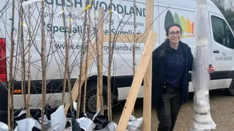 A woman is holding a sapling wrapped in plastic next to several more saplings sitting in buckets. Behind her is a white van that says English Woodlands on the side. 