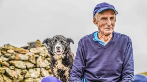 Stephen Wilson A man with white hair and a blue cap sitting next to a wet sheepdog and pile of stones. He is looking to the side wearing a blue jumper. The dog is looking into the camera.