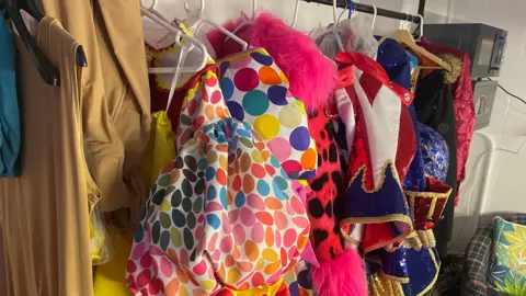 KD Theatre Productions A row of colourful costumes on a rack in a dressing room, on hangers. They have fur and sequins on them, with a chair to the right. There is a lighting box to the right, higher up.