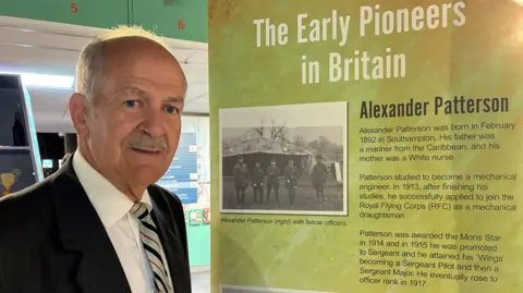 A picture of Rex, who is wearing a dark suit jacket, white shirt and grey striped tie, stood next to a banner that describes Alexander Patterson's professional life.