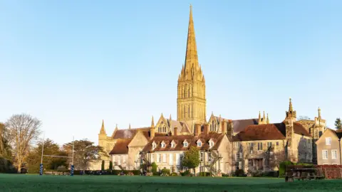 A cathedral spire in the background of several historic stone buildings and a playing field with rugby goal posts at one end