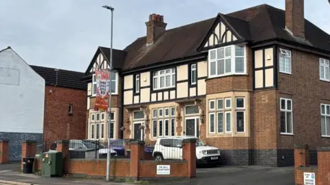 Brian Farmer/BBC A brown brick building decorated on the front with black timber frames. Three cars are parked in front of the building. A sign on a low brown wall in front of the cars says "THE BULL COURTYARD". An orange-and-white banner attached to a lampost in front of the wall says "SHOP SMALL SMILE BIG"