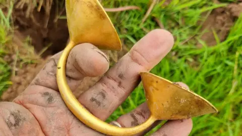 A muddy hand holding a gold dress fastener. The person holding it is standing outdoors in a field with grass and muddy areas.