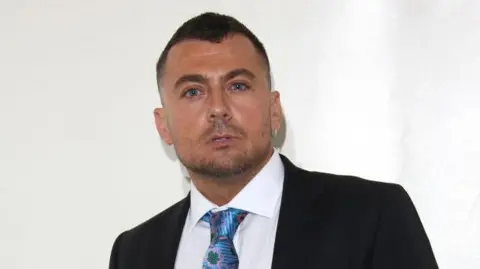 Getty Images Paul Danan, pictured in 2019, wearing a dark suit, white shirt and blue and green tied