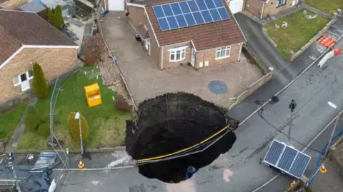 Large sinkhole in street on the drive of one house and into the road, with other homes nearby, barriers in the street.