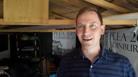 A man in a black and blue striped shirt stands inside a garden shed