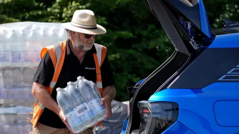 A man in a wide-brimmed hat loads a case of bottled water into the open boot of a car on a sunny day.