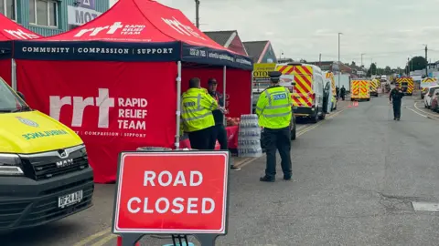 Stephen Huntley/BBC Rapid relief team tents had been put up in Selbourne Road near to the bathroom showroom blaze. A number of emergency services were in attendance including fire, police and ambulance. A red sign with white writing which says ROAD CLOSED has been propped up on the road after a number of roads were partly closed in the area. There are some emergency services personnel standing in the road and within the rapid relief team tent are bottles of water.