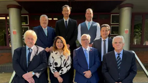 Sandwell Council Seven men and one woman are stood in two rows in front of the Sandwell Council building wearing smart clothes. Councillor Massey is in the back row to the right hand side, wearing a blue tie and grey waistcoat with white shirt.
