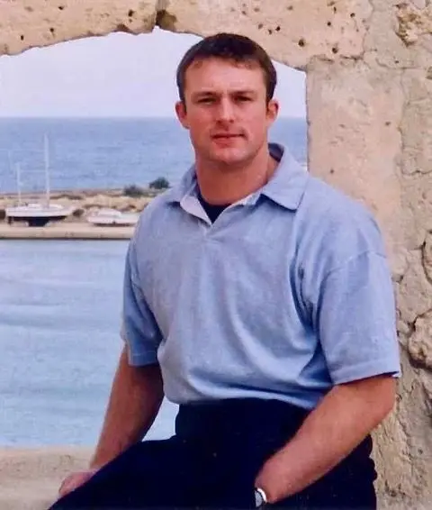 Family photo A man with brown hair, wearing a blue polo shirt and dark trousers. He is sat on a stone ledge with a stone wall and sea visible in the background. He has one hand in his pocket.