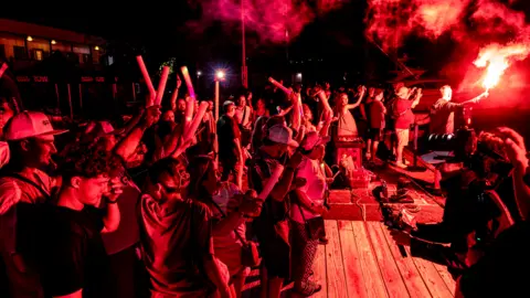 World's Toughest Row A crowd of people cheer and wave as the boat with the four men who made up Team Graft pull into the pontoon. The scene is lit by red flares.