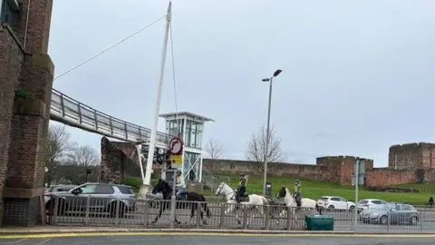 A general view of traffic, including three horses, passing under the Millennium Bridge, with Carlisle Castle in the background.