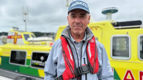 A man stand in the foreground wearing a blue baseball cap with white writing on it, smiling slightly. He's wearing a grey jacket with an orange lifejacket around his neck. A yellow and green marine ambulance is in the background. 