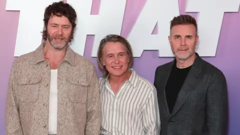 Getty Images Donald wears a white t-shirt underneath a beige jacket. Owen has long hair and wears a white and black stripped shirt. Barlow has a closed grey jacket with a black t-shirt underneath