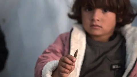 A young child, Razan, holds a large, gold bullet. She is wearing a pink coat with woolly lining and looks forlornly into the camera.