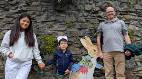 Mark, Lily, and Arthur Lamberton standing in front of a stone wall with a picture of Peter Rabbit. Lily Lamberton has long dark hair and wears a grey hoodie and white trousers. She is holding Arthur's hand. He is a child and has short dark hair and is wearing a paper crown, a blue top and green trousers. Mark Lamberton is bald and wears glasses. He is wearing a grey sweater and camel-coloured trousers. The adults are smiling at the camera, while the boy is looking at it with his mouth open.