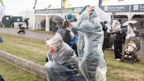 Matt Keeble/PA Wire People in the rain at Royal Cornwall Show. Two people in the foreground have clear plastic ponchos on. People behind them have umbrellas. Heavy rain can be seen. 