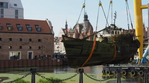 Rotten wooden fishing boat is lifted from a marina by a large industrial crane. In the background are a number of buildings with Gothic architecture